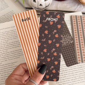 May include: Two rectangular bookmarks with rounded corners. One bookmark features orange and white vertical stripes with the word BACK printed in black. The other bookmark has a dark brown background with orange pumpkins and small leaves with FRONT printed.