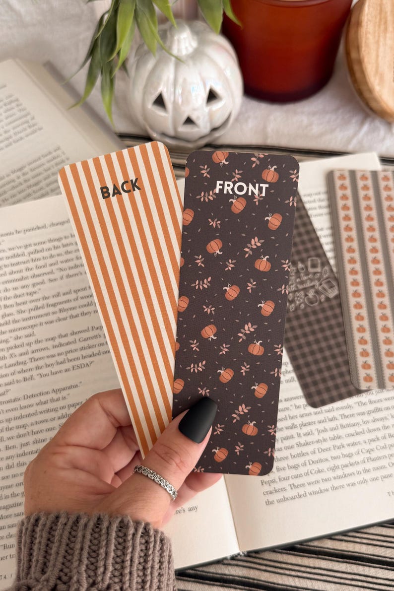 May include: Two rectangular bookmarks with rounded corners. One bookmark features orange and white vertical stripes with the word BACK printed in black. The other bookmark has a dark brown background with orange pumpkins and small leaves with FRONT printed.