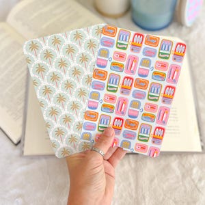 May include: Two decorative book covers are held open, revealing their patterned designs. One features a repeating pattern of palm trees in shades of tan and light green. The other displays colorful illustrations of sardine cans. The covers are open over an open book.