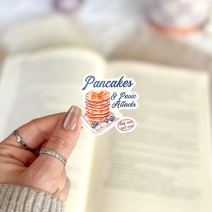 May include: A white sticker with a watercolor illustration of a stack of pancakes with blueberries and a drizzle of syrup. The text "Pancakes & Panic Attacks" is written in blue cursive font. A small red circle with the text "buy me" is in the bottom right corner.