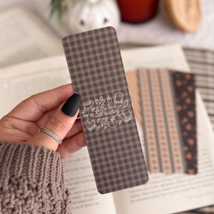 May include: A rectangular bookmark with a dark brown and black gingham pattern. The center features white line art illustrations of a scarf, candle, books, mittens, and an apple, with the text "fall in love with reading" written in script.