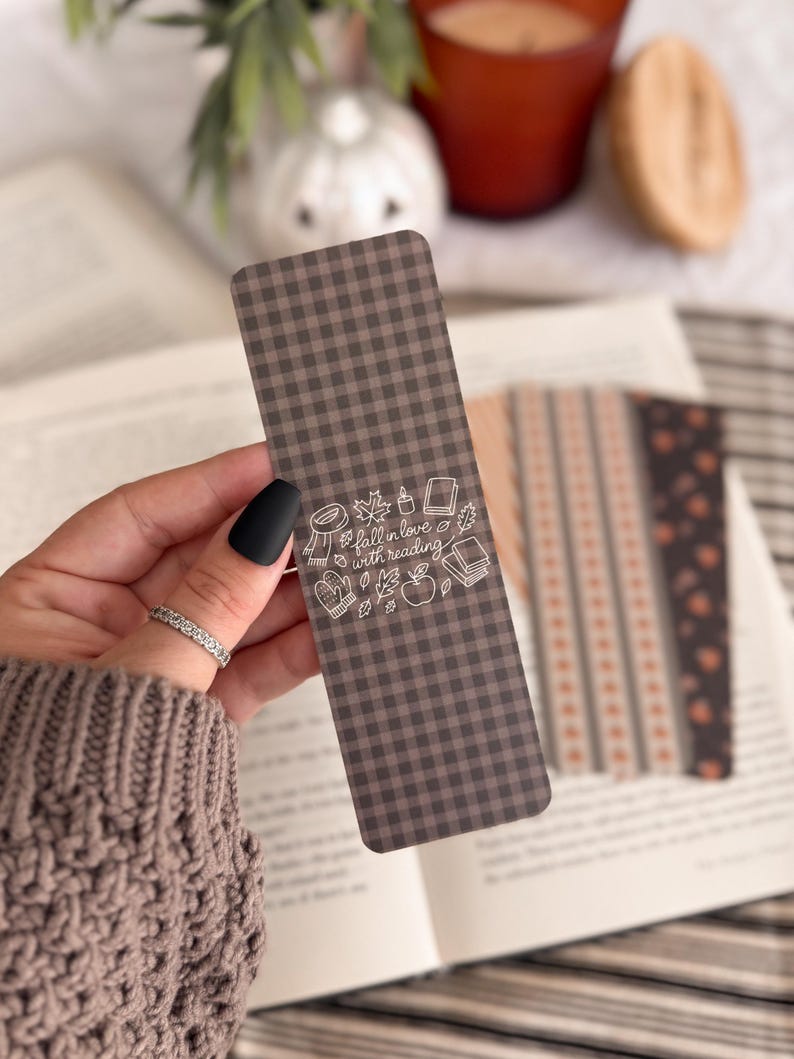 May include: A rectangular bookmark with a dark brown and black gingham pattern. The center features white line art illustrations of a scarf, candle, books, mittens, and an apple, with the text "fall in love with reading" written in script.