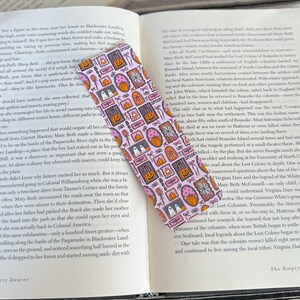 May include: A pink Halloween-themed bookmark with repeating patterns of pumpkins, ghosts, candy corn, and the word "Spooky." The bookmark is rectangular and lies on an open book.