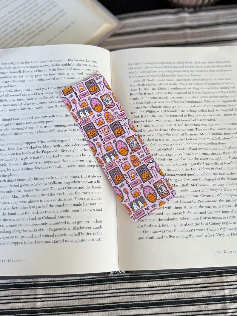 May include: A pink Halloween-themed bookmark with repeating patterns of pumpkins, ghosts, candy corn, and the word "Spooky." The bookmark is rectangular and lies on an open book.