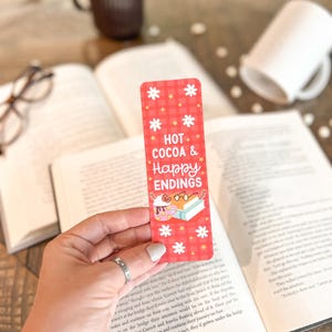 May include: Red and white checkered bookmark with the text "HOT COCOA & Happy ENDINGS" and a cartoon illustration of a person reading a book with a cup of cocoa.