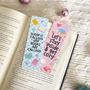 May include: Two colorful bookmarks with winter-themed designs. The first bookmark is blue and white with snowflakes and the text "Snow is Falling and Books are Calling". The second bookmark is pink with snowflakes and the text "Let's Stay Inside & Get Cozy".
