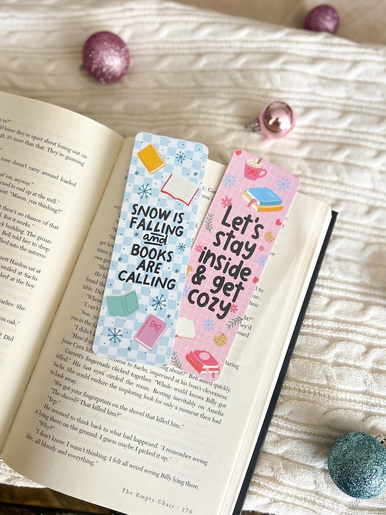 May include: Two colorful bookmarks with winter-themed designs. The first bookmark is blue and white with snowflakes and the text "Snow is Falling and Books are Calling". The second bookmark is pink with snowflakes and the text "Let's Stay Inside & Get Cozy".