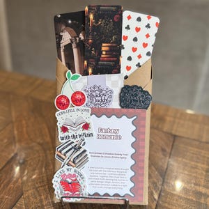 Puede incluir: Un sobre de papel marr&oacute;n con pegatinas y tarjetas decorativas. Las pegatinas incluyen texto como "Oops I fell in love with the villain" y "I like my books". Las tarjetas presentan im&aacute;genes de libros, velas y palos de cartas. Tambi&eacute;n es visible el texto "Fantasy Romance".