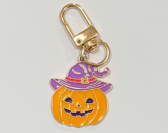 May include: A Halloween-themed keychain featuring an orange pumpkin with a purple witch hat and a gold-colored clasp. The pumpkin has a smiling face with black details. The hat has a red band and a buckle.