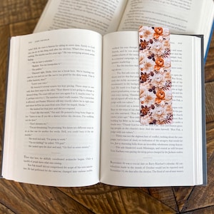 May include: A floral bookmark with orange, white, and brown flowers on a white background. The bookmark is placed on an open book.