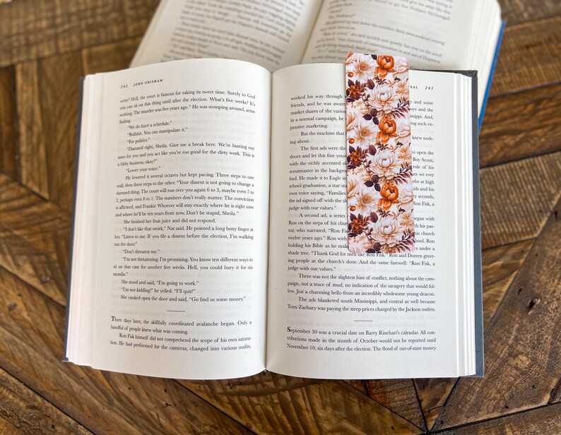 May include: A floral bookmark with orange, white, and brown flowers on a white background. The bookmark is placed on an open book.