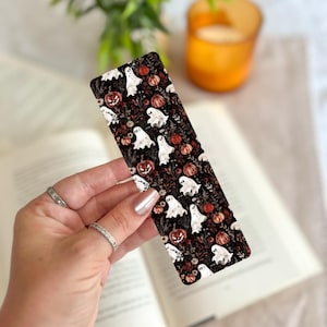 May include: A rectangular Halloween-themed bookmark featuring a repeating pattern of white ghosts, orange pumpkins, and dark foliage on a black background. The bookmark is held over an open book.