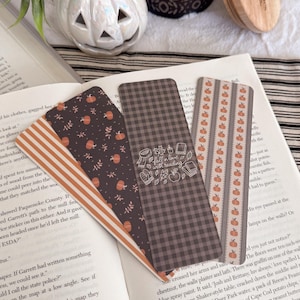 May include: Three rectangular paper bookmarks resting on an open book. One features orange and white stripes, another has a dark background with orange pumpkins and leaves, and the third is a dark grey gingham with the text fall in love with reading.