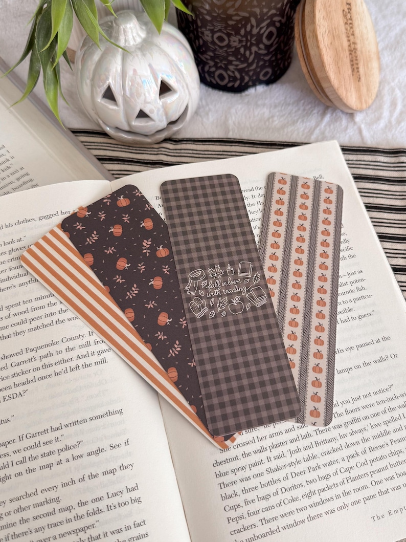 May include: Three rectangular paper bookmarks resting on an open book. One features orange and white stripes, another has a dark background with orange pumpkins and leaves, and the third is a dark grey gingham with the text fall in love with reading.