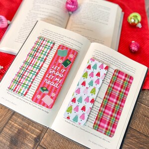 May include: Three Christmas themed bookmarks. One bookmark features a red and white plaid pattern, another has a white background with green and pink Christmas trees, and the third bookmark has a pink and white striped background with the text "Let it snow, let me read".