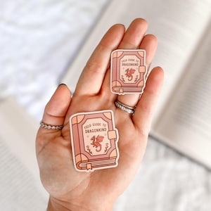 May include: Two pink and white stickers of a closed book with a dragon on the cover. The book is titled "Field Guide to Dragonkind".