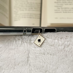 May include: A gold-toned charm in the shape of an Ace of Spades playing card, with a black spade symbol and the letter "A". The charm is attached to a clear phone case, resting on a fluffy white surface.