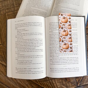 May include: A bookmark with a fall theme featuring a white background with orange pumpkins and brown leaves. The bookmark is made of paper and has a matte finish.