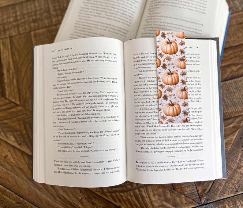 May include: A bookmark with a fall theme featuring a white background with orange pumpkins and brown leaves. The bookmark is made of paper and has a matte finish.