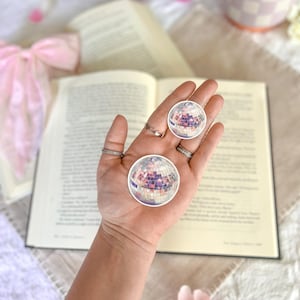 May include: Two silver disco ball stickers on a hand, with a book and flowers in the background.