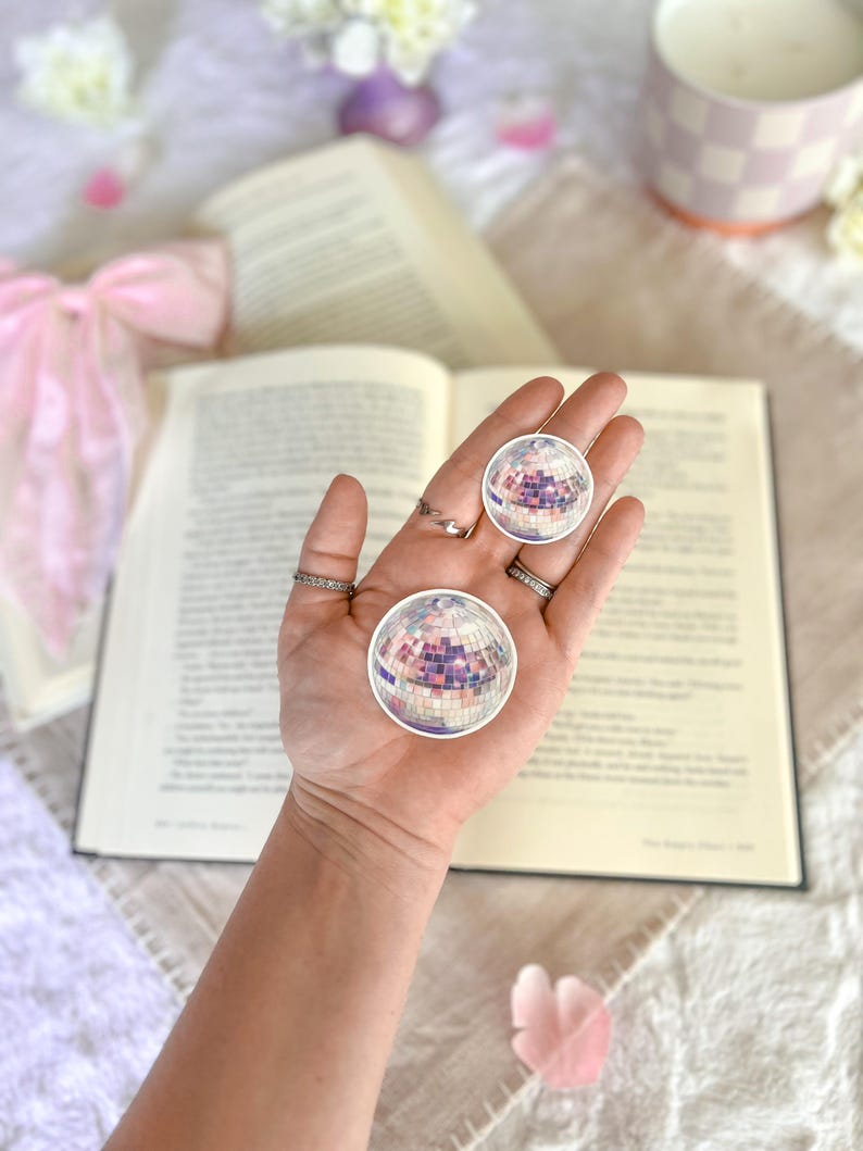 May include: Two silver disco ball stickers on a hand, with a book and flowers in the background.