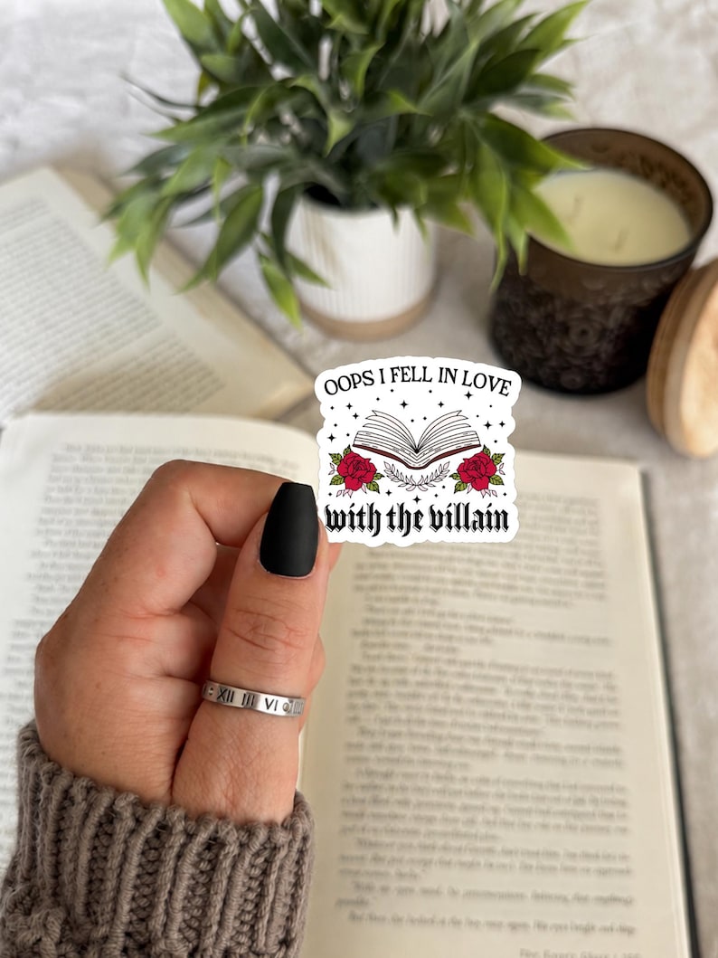 May include: A white die-cut sticker featuring an open book illustration with red roses and the black text "OOPS I FELL IN LOVE with the villain" surrounded by small black stars. The sticker is held against an open book with visible text.
