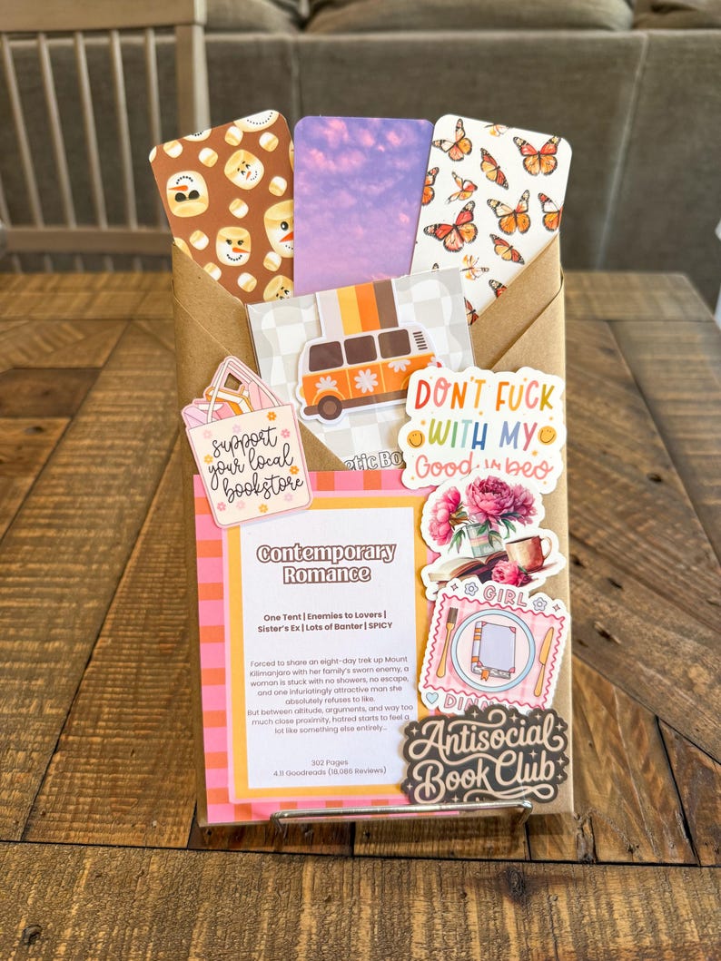 May include: A brown paper envelope containing three bookmarks with snowman, purple sky, and butterfly patterns, decorated with stickers including a van, a bookstore sign, a romance book summary card, floral art, and an Antisocial Book Club label.