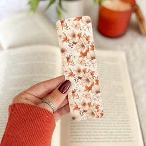 Op de afbeelding: Een rechthoekige boekenlegger met een bloemenpatroon in cr&egrave;me, bruin en roesttinten. De boekenlegger wordt boven een open boek gehouden, met een vage achtergrond met een plant en een kaars.