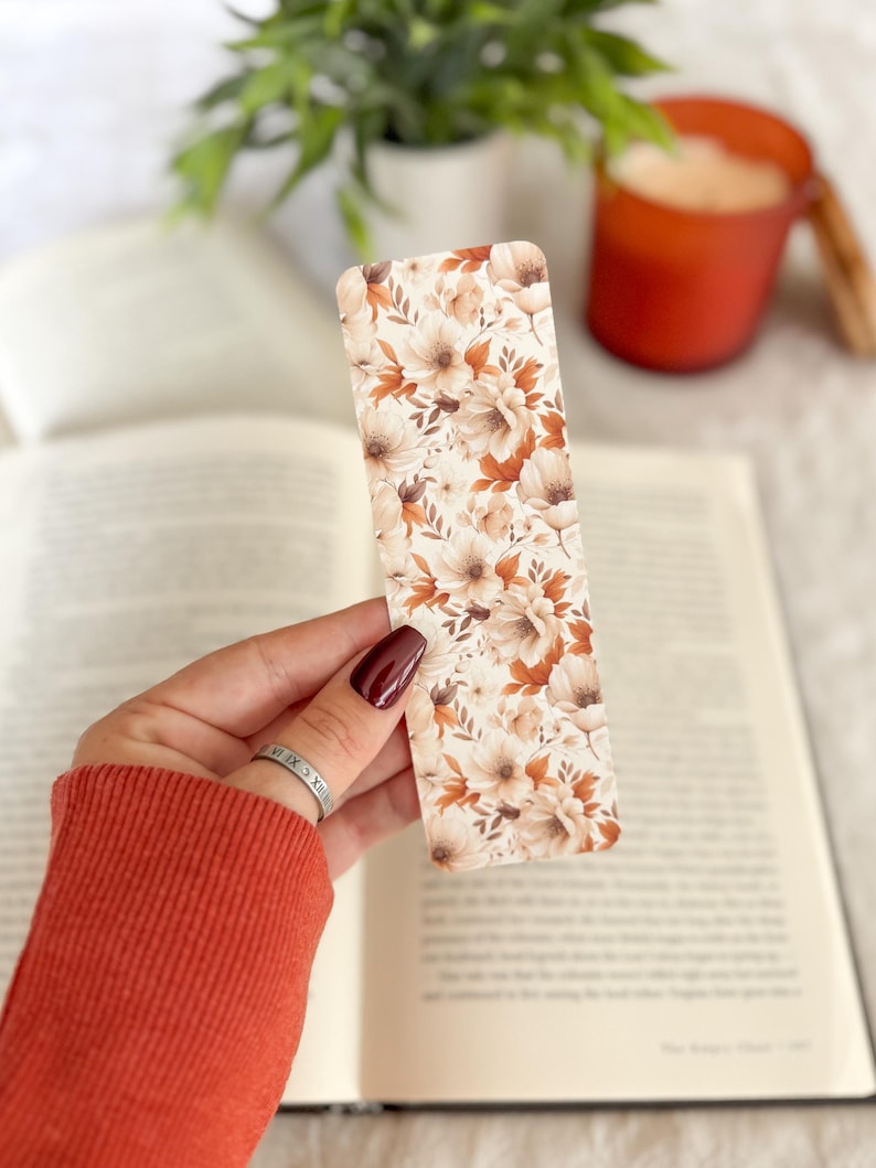 Op de afbeelding: Een rechthoekige boekenlegger met een bloemenpatroon in cr&egrave;me, bruin en roesttinten. De boekenlegger wordt boven een open boek gehouden, met een vage achtergrond met een plant en een kaars.