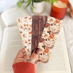 Op de afbeelding: Drie decoratieve bladwijzers met bloemen- en houtnerfdesigns. De bladwijzers hebben cr&egrave;me, bruine en oranje kleuren. De bladwijzers worden voor een open boek gehouden, met een kaars en een plant op de achtergrond.