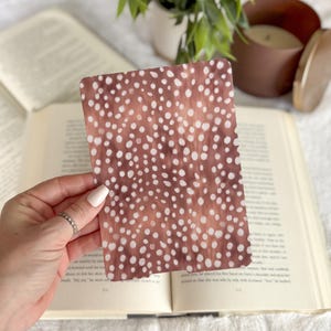 May include: A rectangular card with a brown background and white polka dots is held over an open book. The card has rounded corners and a textured appearance. The image also includes a plant, a candle, and a book.