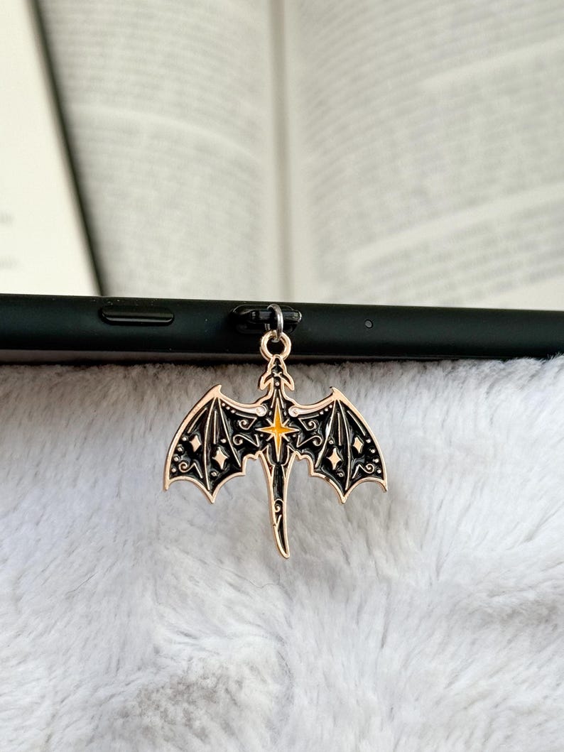 May include: A gold-colored dragon charm with black detailing and a yellow starburst center. The dragon has outstretched wings with intricate patterns and is attached to a black phone. The charm is set against a soft, white background.