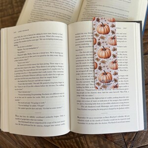 May include: A close-up of a bookmark with a fall theme. The bookmark features a white background with orange pumpkins, brown leaves, and a small tag that says "Little Shoppe".