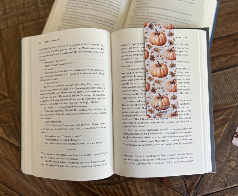 May include: A close-up of a bookmark with a fall theme. The bookmark features a white background with orange pumpkins, brown leaves, and a small tag that says "Little Shoppe".