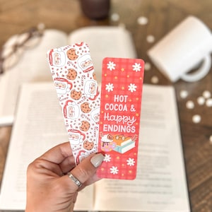 May include: Two Christmas-themed bookmarks. One bookmark features a red and white checkered pattern with white flowers and the text "Hot Cocoa & Happy Endings". The other bookmark features a white background with a repeating pattern of Santa Claus, candy canes, and chocolate chip cookies.