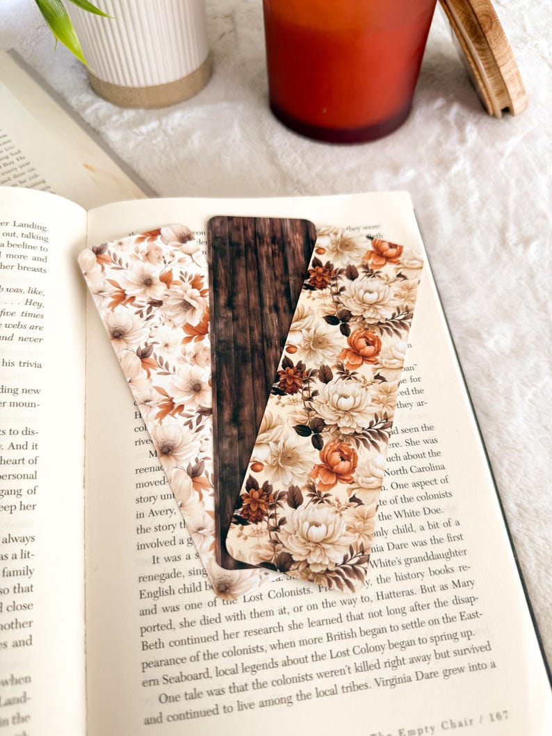 Op de afbeelding: Drie decoratieve bladwijzers met bloemen- en houtnerfdesigns liggen op een open boek. De bladwijzers hebben een mix van cr&egrave;me, bruine en oranje bloempatronen en een centrale bladwijzer met een houtnerftextuur.