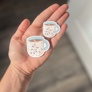 May include: Two stickers of a white mug with a blue rim and handle. The mug is filled with a brown liquid and has a string of colorful lights wrapped around it.