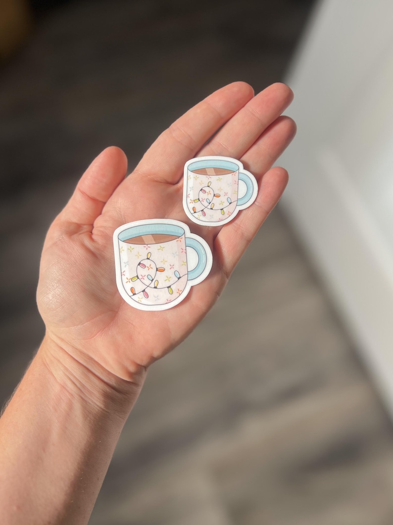 May include: Two stickers of a white mug with a blue rim and handle. The mug is filled with a brown liquid and has a string of colorful lights wrapped around it.