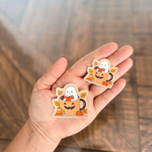 May include: Two wooden Halloween-themed stickers. Each sticker features a white ghost reading a book inside a jack-o'-lantern mug, surrounded by fall leaves and a small pumpkin. The stickers are on a wooden surface.