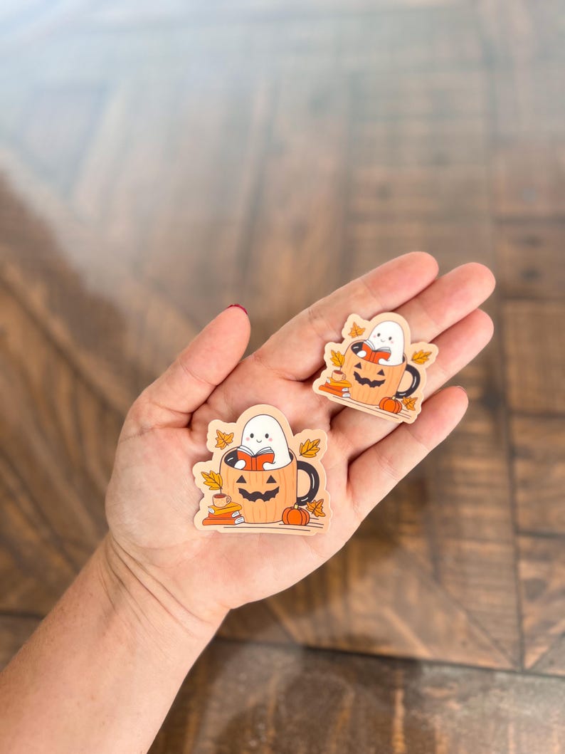 May include: Two wooden Halloween-themed stickers. Each sticker features a white ghost reading a book inside a jack-o'-lantern mug, surrounded by fall leaves and a small pumpkin. The stickers are on a wooden surface.