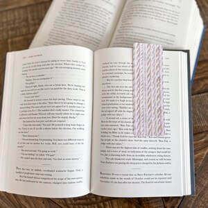 May include: A close-up of an open book with a pink knitted bookmark. The book is open to a page with text about a trial. The bookmark is made of a soft, textured yarn.