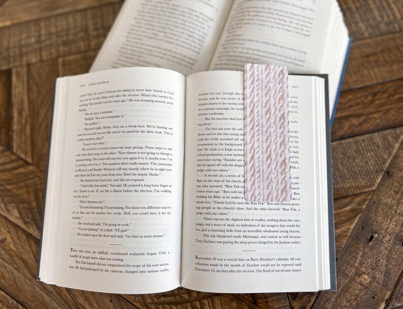 May include: A close-up of an open book with a pink knitted bookmark. The book is open to a page with text about a trial. The bookmark is made of a soft, textured yarn.