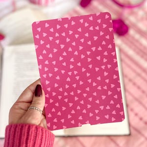 May include: A pink bookmark with a pattern of small white hearts. The bookmark is being held by a hand.