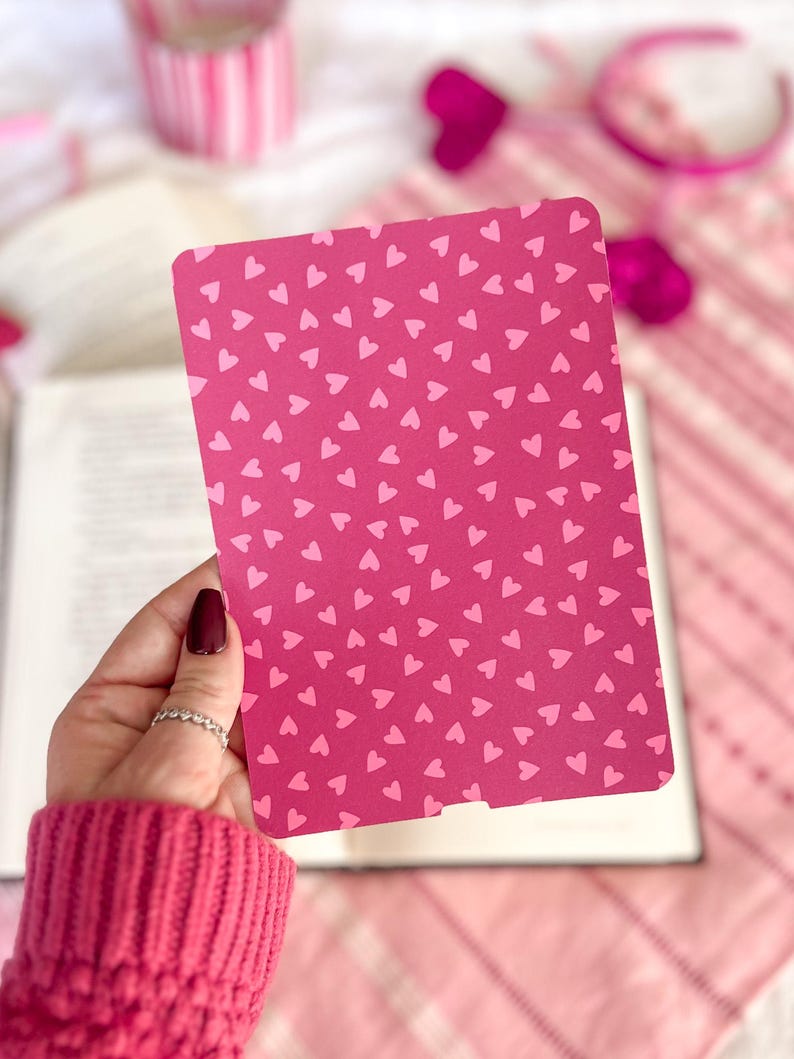 May include: A pink bookmark with a pattern of small white hearts. The bookmark is being held by a hand.