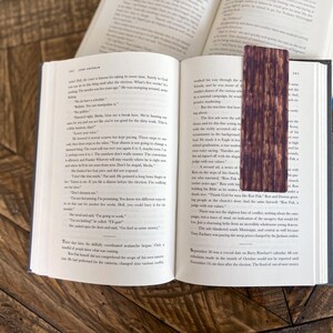 May include: A close-up of an open book with a brown wooden bookmark. The bookmark has a wood grain pattern and is slightly darker in color than the book pages. The text on the book pages is visible, but the words are not clear enough to read.