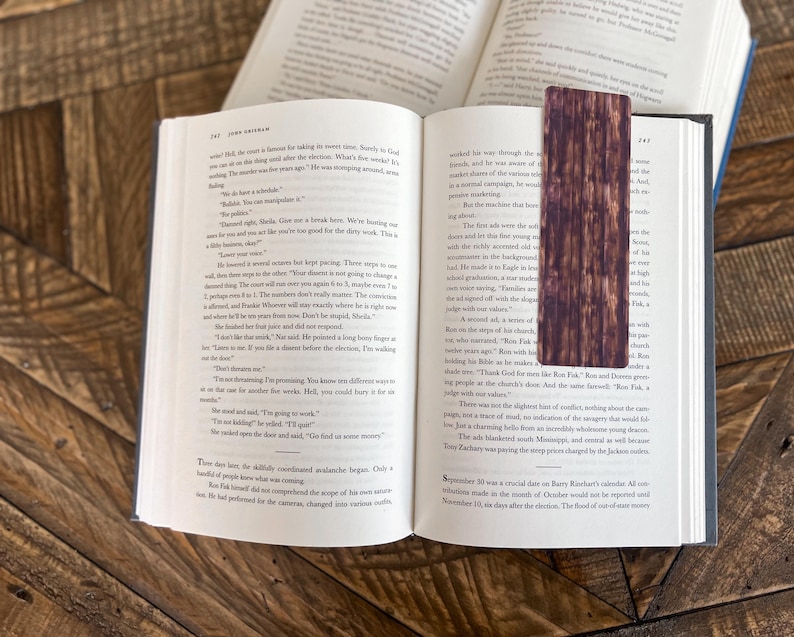 May include: A close-up of an open book with a brown wooden bookmark. The bookmark has a wood grain pattern and is slightly darker in color than the book pages. The text on the book pages is visible, but the words are not clear enough to read.
