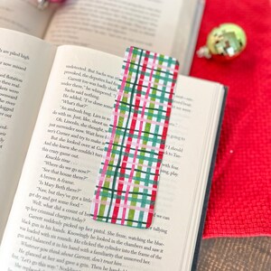 May include: A red, green, and white plaid bookmark with a pink stripe. The bookmark is lying on top of an open book with text about an ambush.