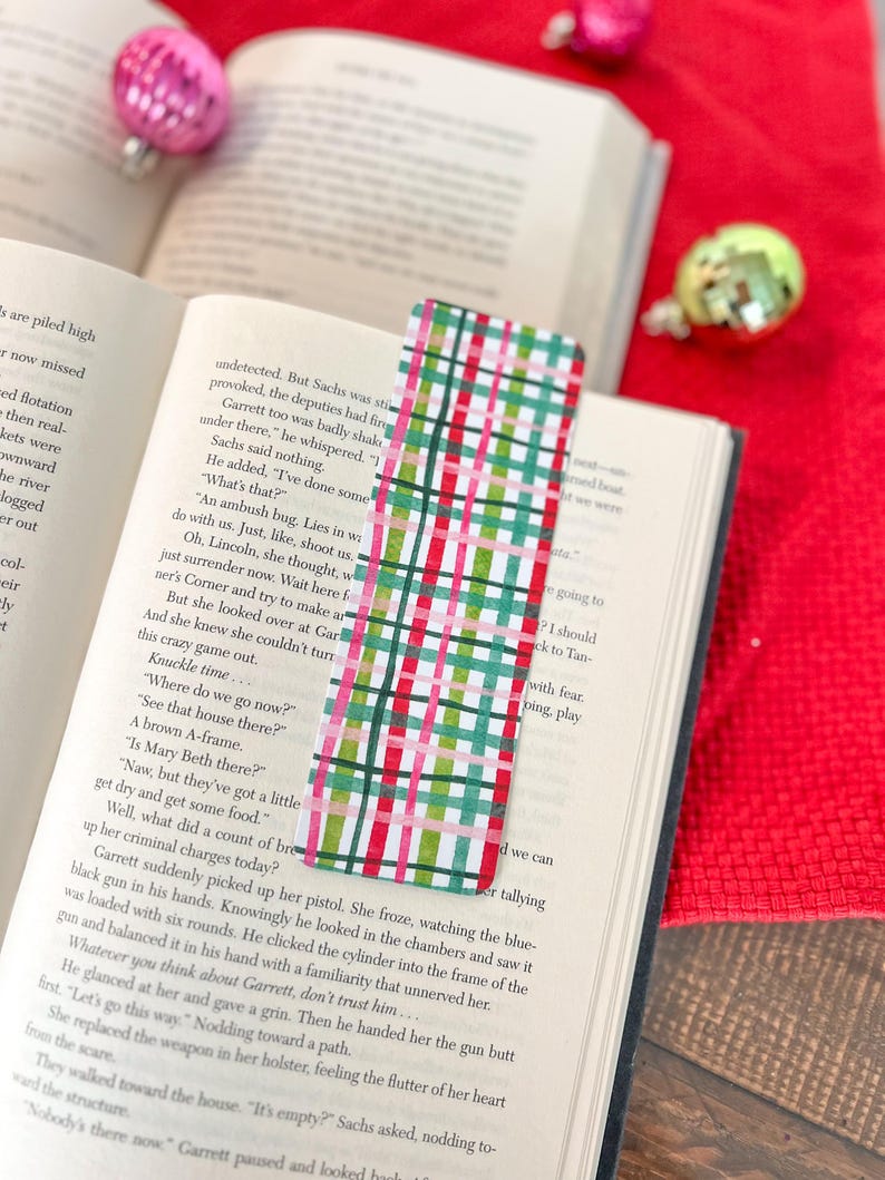 May include: A red, green, and white plaid bookmark with a pink stripe. The bookmark is lying on top of an open book with text about an ambush.