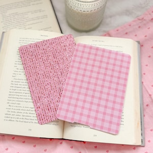 May include: Two rectangular bookmarks resting on an open book. One bookmark features a pink and white gingham check pattern, while the other displays a textured pink surface with vertical rows of small, shimmering copper-colored glitter accents.