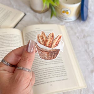 May include: A sticker featuring a watercolor illustration of several baguettes in a woven basket lined with white cloth. The baguettes are golden brown with a textured appearance. The basket is brown and the sticker is held over an open book.